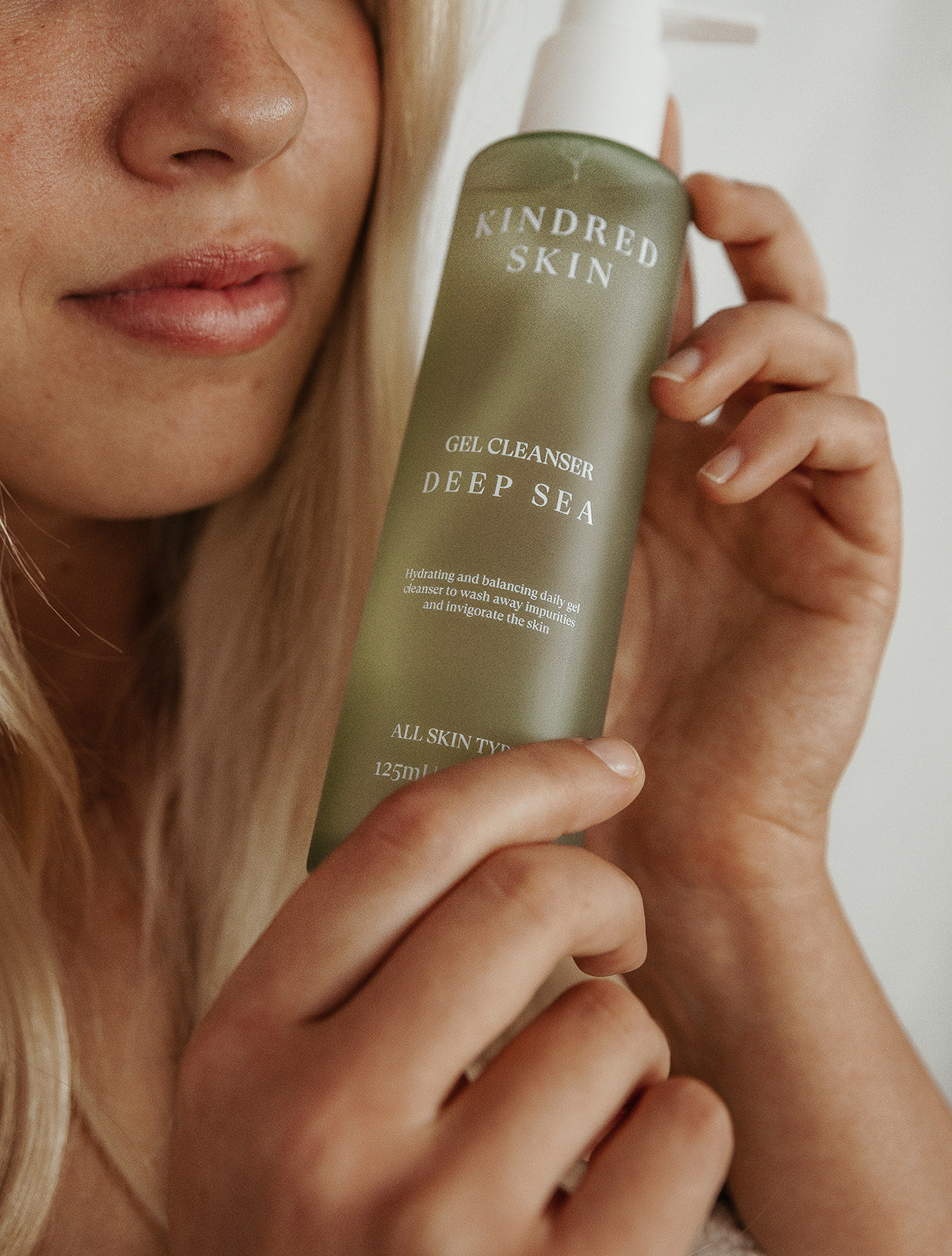 blonde pretty girl with no acne holding a green kindred skincare deep sea gel cleanser bottle with a bamboo bath towel